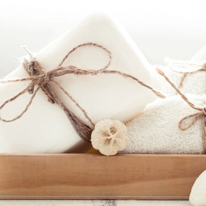 Weißes Handtuchset mit dekorativer Kordel auf einem Holztablett, daneben weiße Naturseife und Bimssteine.