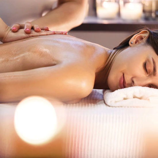 Eine Frau liegt entspannt auf einer Massageliege, während ihr Rücken von einer Masseurin sanft eingeölt und massiert wird. Ihr Kopf ruht auf einem weißen Handtuch, die Augen sind geschlossen. Im Hintergrund sorgen Kerzen für eine stimmungsvolle Wellness-Atmosphäre.